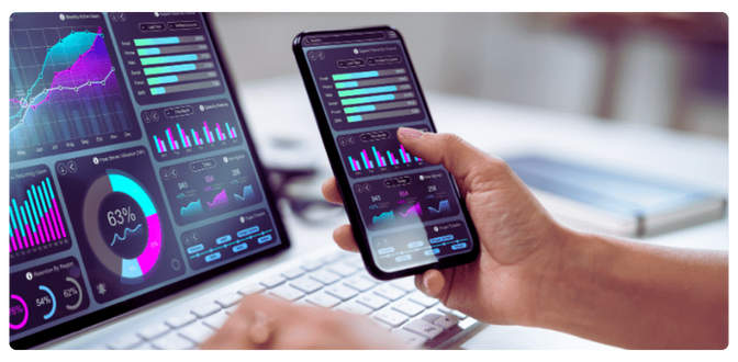 A person holding a mobile device with charts and graphs showing data from website performance. They're comparing the data to that which is shown on a laptop showing sales data. 